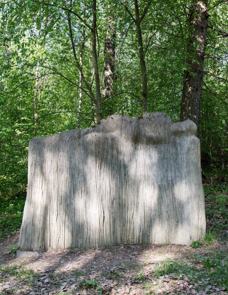 Puistoon sijoitettu betoninen veistos, joka kuvaa pajukorokkeesta kasvavaa, lepäävää naisfiguuria.