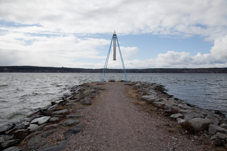 Geometrinen, avointa kolmiota muistuttava neljän metallipuomin varassa seisova taideteos, jonka keskellä yläosasta roikkuu metalliputkista toteutettu tuulikello.