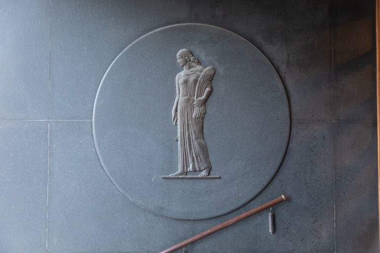 Reliefi, jossa pitkään, toogamaiseen vaatteeseen puettu paljasjalkainen naishahmo pitää kainalossaan viljalyhdettä.