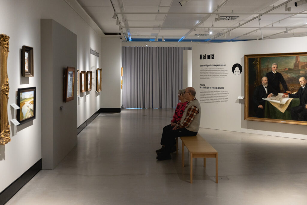 Kaksi henkilöä istuu penkillä ja katsoo maalausta Juuret-näyttelyssä.