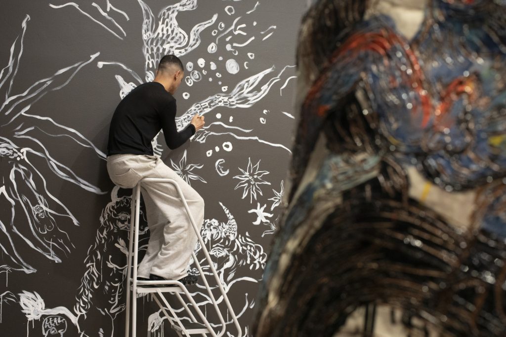 Karim Boumjimar maalaamassa seinämaalausta valkoisella maalilla mustaa seinää Malvan näyttelysalissa.