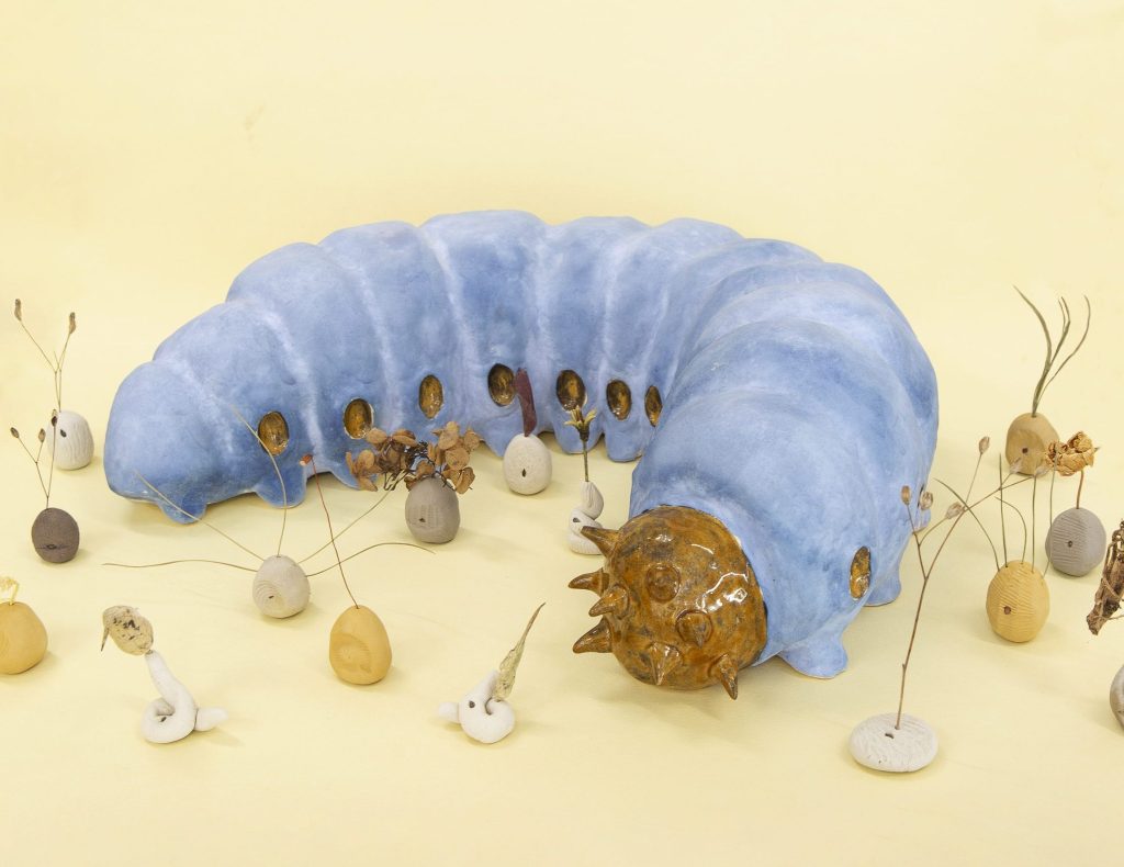 A ceramic blue and yellow caterpillar surrounded by small round ceramic elements, from which plant sprouts emerge.
