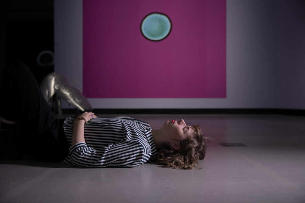 Henkilö makaa maassa silmät kiinni taideteoksen edessä.