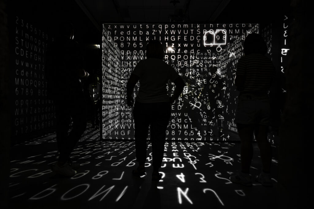 Henkilö seisoo hämärässä näyttelysalissa ja katselee ympäri salia heijastettuja valkoisia numeroita ja kirjaimia.